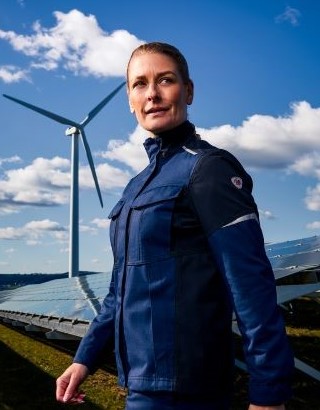Eine Frau trägt Multinorm Schutzkleidung. Im Hintergrund ist eine Windkraftanlage und Solaranlagen zu sehen.